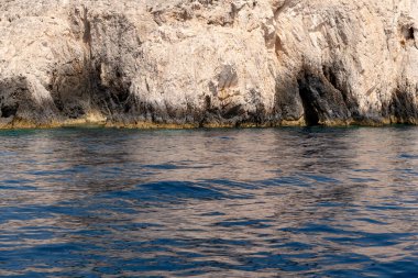 Engebeli bir kireçtaşı kayalığı, yumuşak güneş ışığı altında bir Yunan adasının kıyı şeridi boyunca sakin mavi denizle buluşuyor..