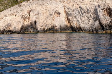 Engebeli bir kireçtaşı kayalığı, yumuşak güneş ışığı altında bir Yunan adasının kıyı şeridi boyunca sakin mavi denizle buluşuyor..