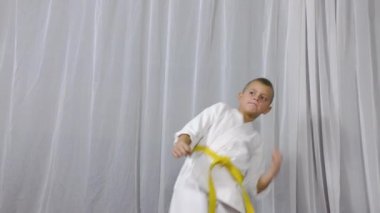 On a light background, an athlete in karategi and with a yellow belt performs kicks