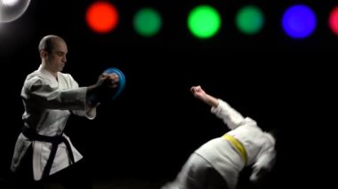 Sarı kuşaklı trenleri olan erkek sporcu siyah arka planda renk değiştiren spot ışıklarıyla simülatöre tekme atıyor.