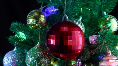 Koyu kırmızı kareli bir Noel topu, toplar ve çelenklerle süslenmiş Noel ağacı dallarının arka planında dönüyor.