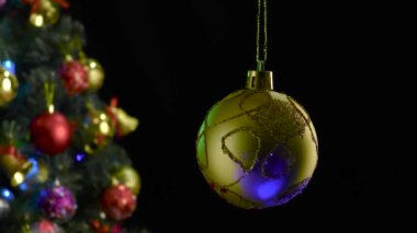 Siyah bir arka planda, süslenmiş bir Noel ağacının yanında dönen sarı bir Noel topu.