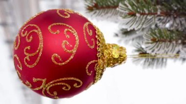 Yarı bulanık bir arkaplanda, bir Noel ağacının karla kaplı dallarında asılı duran kırmızı bir Noel topu.