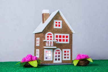 Penceresinde kırmızı ışık olan kahverengi bir oyuncak evin yanında çiçekler var.