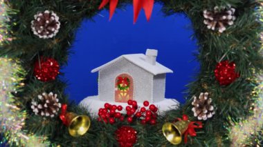 Beyaz bir ev mavi arka planda süslü bir Noel çelenginin ortasında dönüyor.