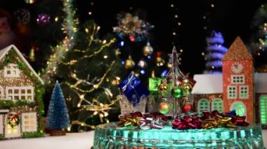 Küçük cam bir Noel ağacı ve renkli hediyeler evlerin ve Noel ağaçlarının arka planında dönüyor.