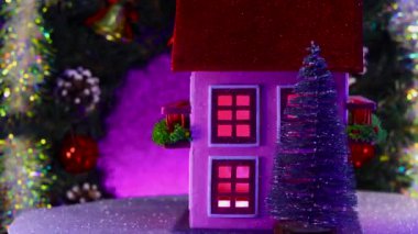 Noel ağaçlarıyla çevrili pembe iki katlı bir ev süslü bir Noel çelenginin arka planında dönüyor.