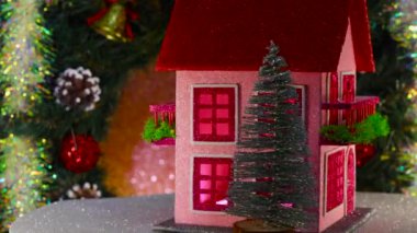 Noel ağaçlarıyla çevrili pembe dekore edilmiş iki katlı bir ev bir Noel çelenginin arka planında dönüyor.