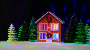 Pencerelerinde çok renkli ışıklar olan iki katlı bir Noel evi Noel çelengi ve Noel ağaçları arasında uçan animasyonla süslenmiş.