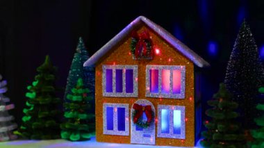 Pencerelerinde çok renkli ışıklar olan iki katlı bir Noel evi Noel çelengi ve Noel ağaçları arasında uçan animasyonla süslenmiş.