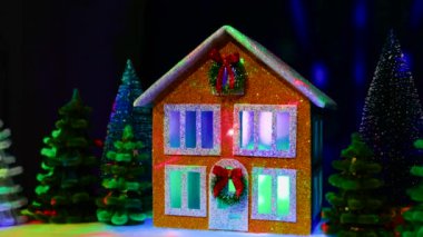 Pencerelerinde çok renkli ışıklar olan iki katlı bir Noel evi Noel çelengi ve çok renkli Noel ağaçları arasında uçan animasyonla süslenmiş.