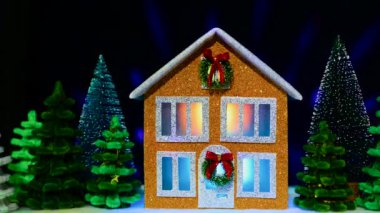 İki katlı kahverengi bir ev, çok renkli köknar ağaçları arasında iki Noel çelengiyle süslenmiş. Koyu mavi arka planda, uçan ışık huzmeleri var.