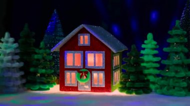 Kırmızı ve yeşil iki katlı ev, koyu mavi arka plandaki çok renkli Noel ağaçları arasında Noel çelengi ve uçan animasyonla süslenmiş.