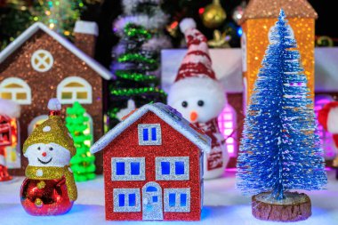 Noel kasabasının ön planında, camlarında mavi ışıklar olan, Noel ağacı ve Noel ağacı süslemeleri olan kırmızı bir ev var..