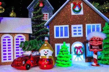 Pencerelerinde mavi ve mor ışıklar olan Noel evlerinin yanında Noel ağacı süslemeli kırmızı bir araba var. Kardan adam ve Noel ağacı şeklinde.