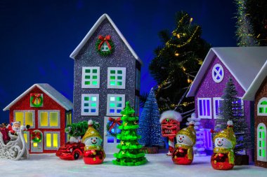 Noel köyünün merkezinde, pencerelerinde Noel ağacı olan evler, kızağında Noel Baba olan kırmızı bir araba ve kardan adam şeklinde Noel ağacı oyuncakları.