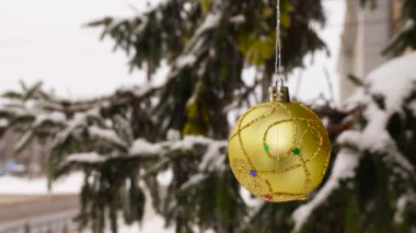 Sarı bir Noel topu, yarı bulanık arka planda, karla kaplı dalların yanında ağırdır.