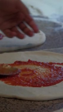 Şefin ellerinin taze pizza hamuruna domates sosu sürerken çekilmiş yakın çekim görüntüleri. Profesyonel bir mutfakta pizza hazırlama süreci. Otantik İtalyan mutfağı yemek pişirme aşamasında yakalanır.