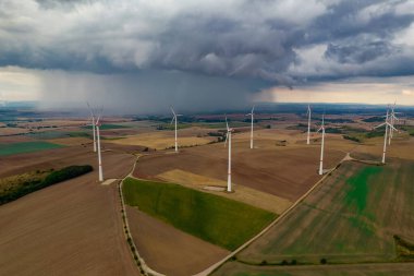 İHA uçuşu, çok sayıda rüzgar türbininin döndüğü büyük bir alanın üzerinde yüksek irtifada. Rüzgar türbinleri uzun bir gölge bırakıyor çünkü güneş batıyor. Arka planda, fırtına gökyüzünde.