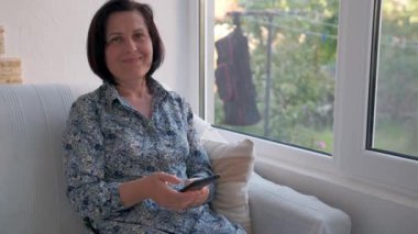 Zarif bir yetişkin kadın verandada oturuyor ve telefonundaki video bağlantısıyla konuşuyor.