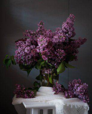 Masadaki beyaz bir vazoda leylak çiçeklerinin güzel bir düzeni sergilenir. Çiçekler tomurcuklanıyor ve canlı renkleri canlılık ve canlılık hissi yaratıyor..