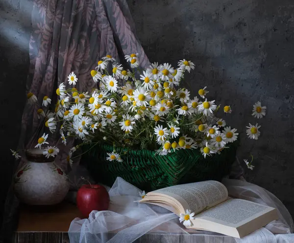 Kırmızı bir elma ve açık bir kitapla kaliteli bir sürahi içinde neşeli bir papatya koleksiyonu, büyüleyici bir sahne yaratıyor.