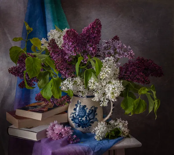 Kitapların ve renkli kumaşın yanına kokulu bir sürahiye yerleştirilmiş leylakların hoş ve büyüleyici bir düzeni.