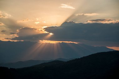 Bulgaristan 'da gün batımından önce bulutların arasından parlayan parlak güneş ışığı, dağ sırasının karanlık sahnesine yansıyor..
