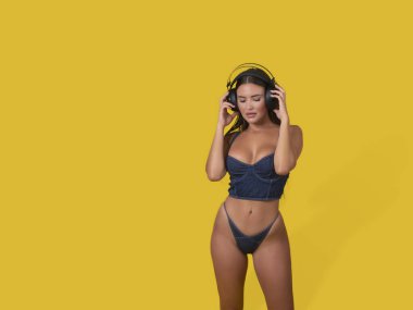 Büstiyer sutyen ve külot giyen şehvetli bir kadın model kablosuz kulaklıkları ayarlıyor ve sarı arka planda kapalı gözlerle favori müziğin tadını çıkarıyor.