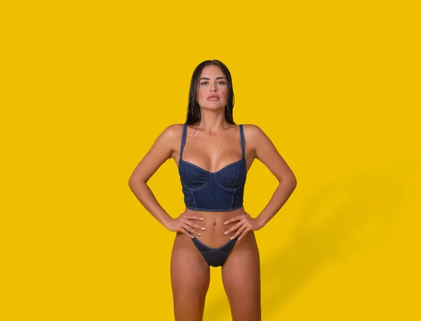 Self assured woman in denim bustier bra and panties holding hands on waist and looking at camera against yellow background