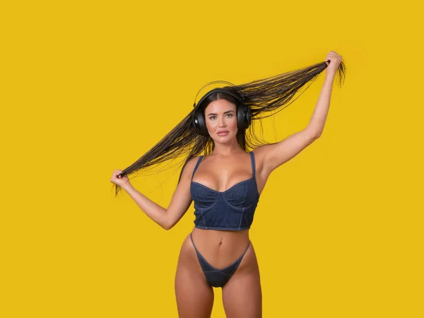 Female model in underwear listening to music in wireless headphones and touching long hair against yellow background