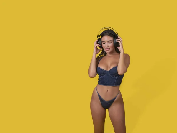 穿着紧身胸衣和内裤的性感女模特 在黄色背景下调整无线耳机 欣赏最喜欢的闭眼音乐 图库图片