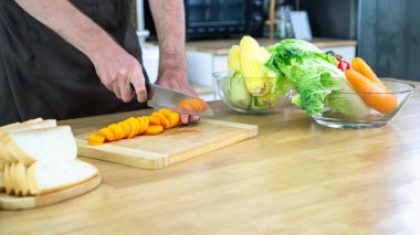 Sağlıklı sebzeler ve ekmeklerle çevrili modern bir mutfağın ahşap kesme tahtasıyla taze havuç doğrayan bir adamın yakın plan fotoğrafı.