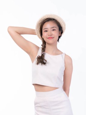 Saçları örgülü, saman şapka takan ve özgüvenle gülümseyen, elleri başının arkasında, beyaz arka planda izole bir şekilde sıradan bir poz veren, açık renk bir yaz elbisesi içinde güzel bir Asyalı kadın.