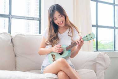 Uzun kahverengi saçlı genç bir Asyalı kadın, kanepede oturuyor ve açık mavi bir ukulele çalıyor.
