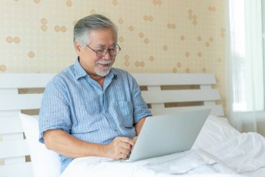 Mutlu Asyalı son sınıf öğrencisi evde yatağında dinlenirken dizüstü bilgisayar kullanıyor, yaşlı erkeklerin internette geziniyor ya da yatak odasında, yaşam tarzında ve teknolojide yaşlılar için çalışıyor.