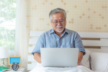 Mutlu üst düzey Asyalı adam rahat yatak odasında dizüstü bilgisayar kullanıyor, gözlük takıyor ve günlük giysiler giyiyor, evden çalışıyor veya internette geziniyor, evde yaşlılar yaşam tarzı ve dijital teknoloji kavramı