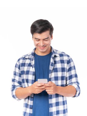 Mutlu Asyalı genç adam gülümserken akıllı telefon kullanıyor, günlük mavi kareli gömlek giyiyor, beyaz bir arka plan, iletişim ve teknoloji kavramında izole edilmiş ve telefonu tutuyor.