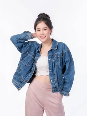Güzel, genç Asyalı bir kadın, kot ceket ve kesim bluzuyla güvenle gülümsüyor, eli boynun arkasında, şık ve çekici bir poz, sıradan moda görünüşü, beyaz arka planda izole edilmiş.