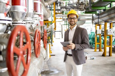 Power plant engineer standing by gas pipeline inside energy production factory and checking results on digital tablet.