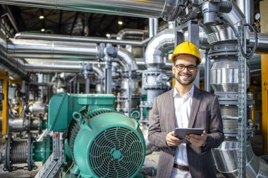 Portrait of power plant engineer standing by gas engines inside energy production factory and checking results on digital tablet.