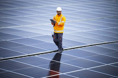 Professional worker using digital tablet and walking on rooftop checking solar panel installations and energy production.