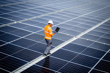 Renewable energy engineer walking through large solar power plant and checking electricity production on his laptop computer. Sustainable energy source and eco friendly technologies.