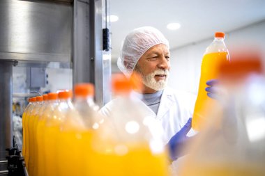 Experienced factory worker holding PET bottle and doing visual inspection of orange juice in bottling plant.