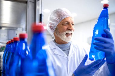 Experienced food factory supervisor in white sterile uniform holding PET bottle and doing visual inspection of water in bottling plant.