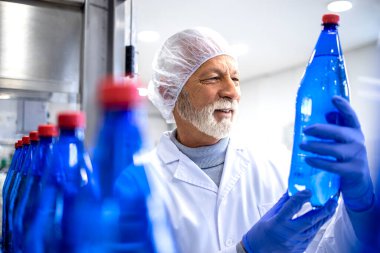 Experienced factory worker holding PET bottle and doing visual inspection of water in bottling plant.
