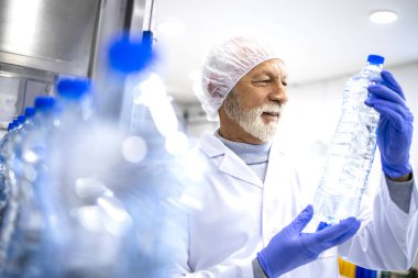 Experienced factory worker holding PET bottle and doing visual inspection of water in bottling plant.
