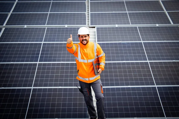 Portrait view of professional worker contractor holding thumbs up and standing in solar energy power plant after successful installation of photovoltaic cell panels.
