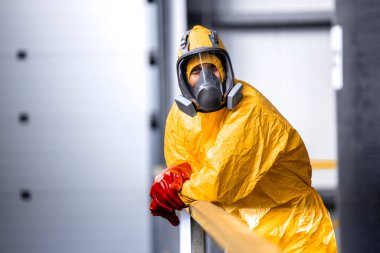 Portrait of factory worker in protective yellow suit, gas mask and gloves leaning against the fence inside chemicals production plant.