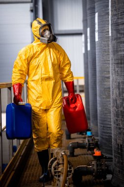 Experienced worker in yellow protection suit and gas mask working in chemicals factory and walking by large acid tanks.